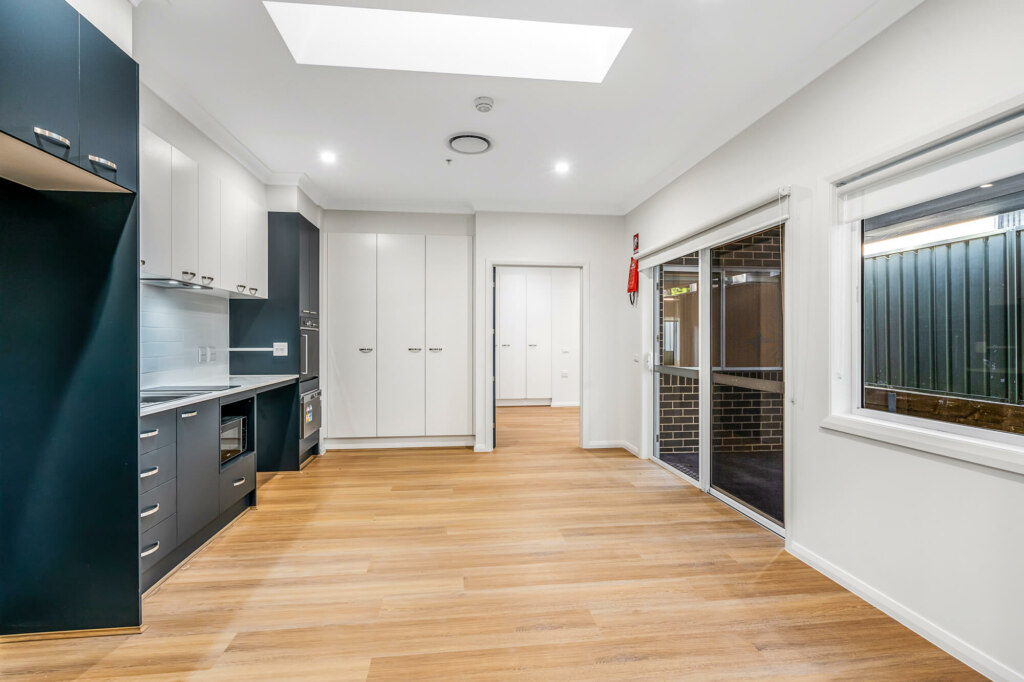 Modern kitchen with dark cabinetry, light countertops, and wooden flooring. The room features a large window, sliding door, ample storage, and recessed lighting. Located in the picturesque Sutherland Shire of Sydney, a doorway leads to another room.