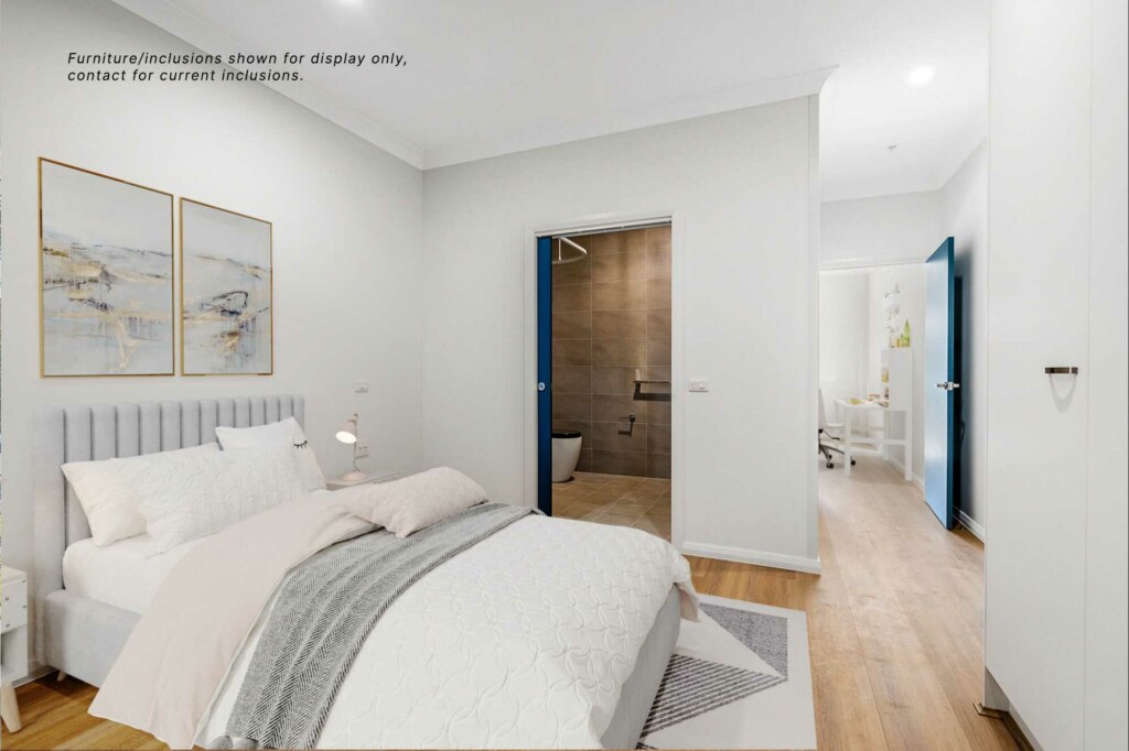 A modern bedroom in Sutherland Shire, featuring a neatly made bed, two abstract paintings on the wall, and an en-suite bathroom. The room leads to a hallway with a visible study area in the distance. Furniture/inclusions shown for display only; contact for current inclusions.