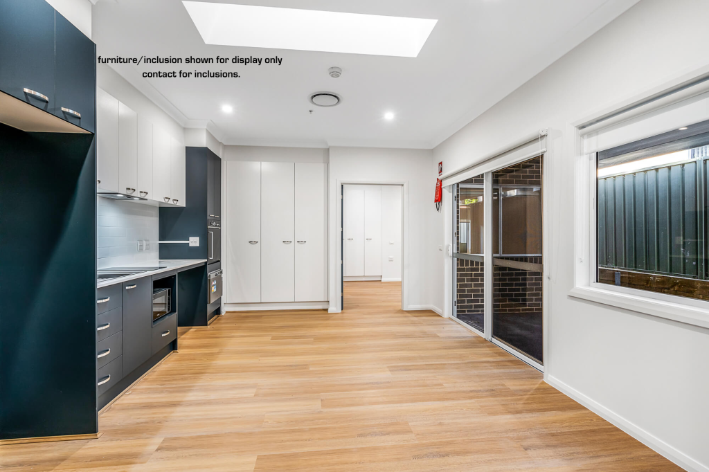 Modern kitchen and living area in C Wamberal with wood flooring, white cabinets, built-in appliances, large windows, and sliding glass door to outdoor space; inclusion note visible on wall.