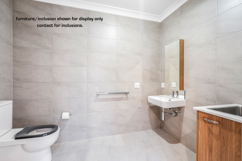 Accessible bathroom with tiled walls, a toilet, wall-mounted sink, mirror, and a small counter with a cabinet. Text on the wall notes furniture/inclusion display. Located in Narraweena on Sydney’s Northern Beaches.