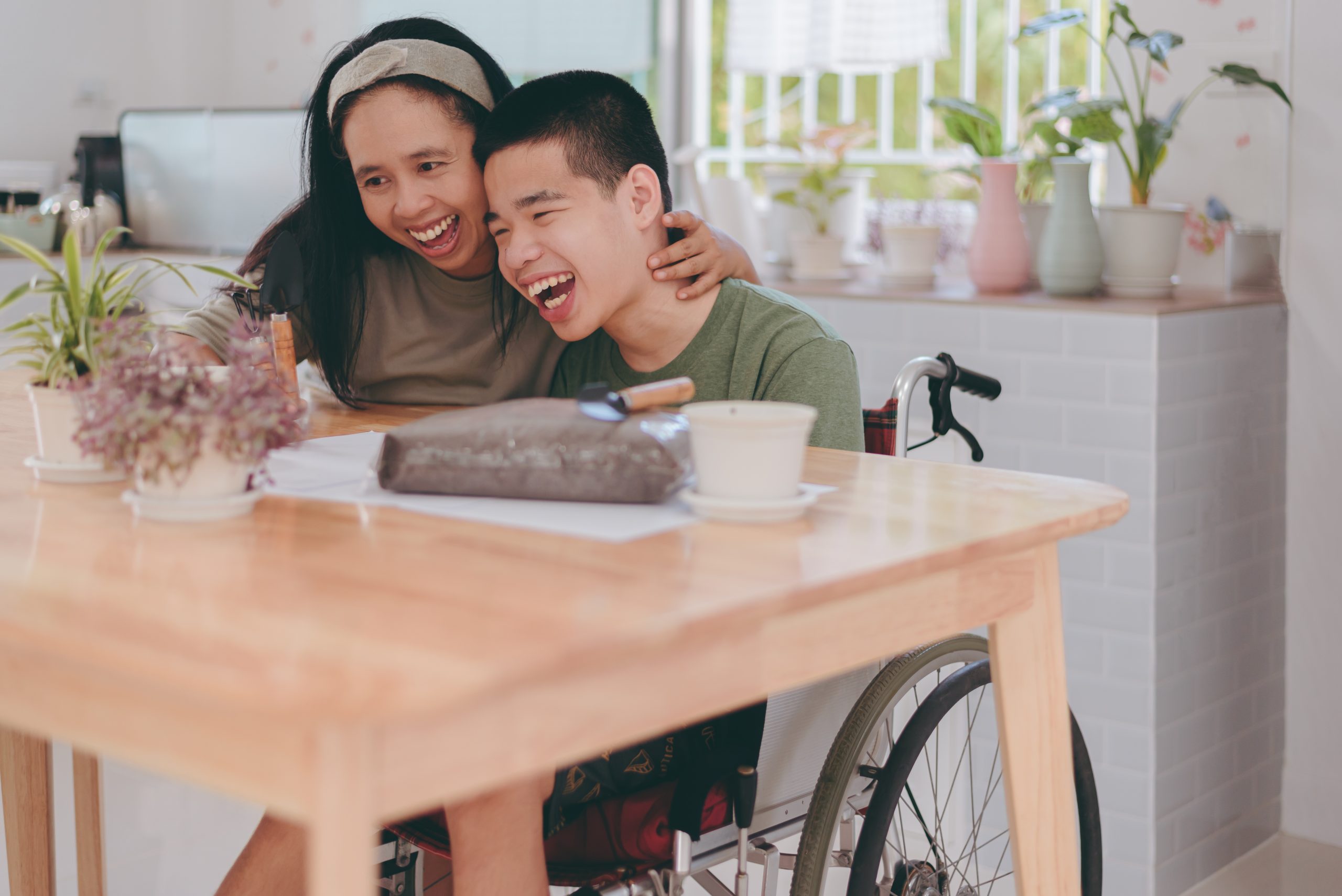 A woman hugs a smiling young man in a wheelchair at a kitchen table with a chocolate cake and potted plants.