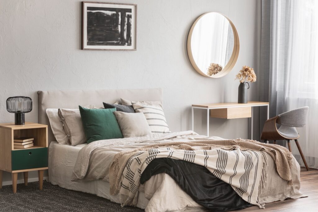 Located in North Gosford, this modern bedroom features a neatly made bed accented with green and beige pillows. A bedside table with a lamp, a round mirror above a wooden desk, and a large window with curtains on the right complete the inviting space.