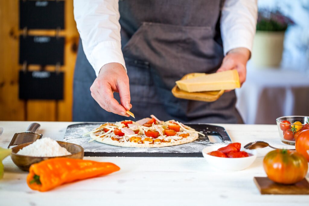 In a cozy North Gosford kitchen, a person in a gray apron prepares pizza, skillfully adding fresh ingredients to the dough base. Vegetables and grated cheese are spread across the table, ready to transform into a delicious masterpiece.