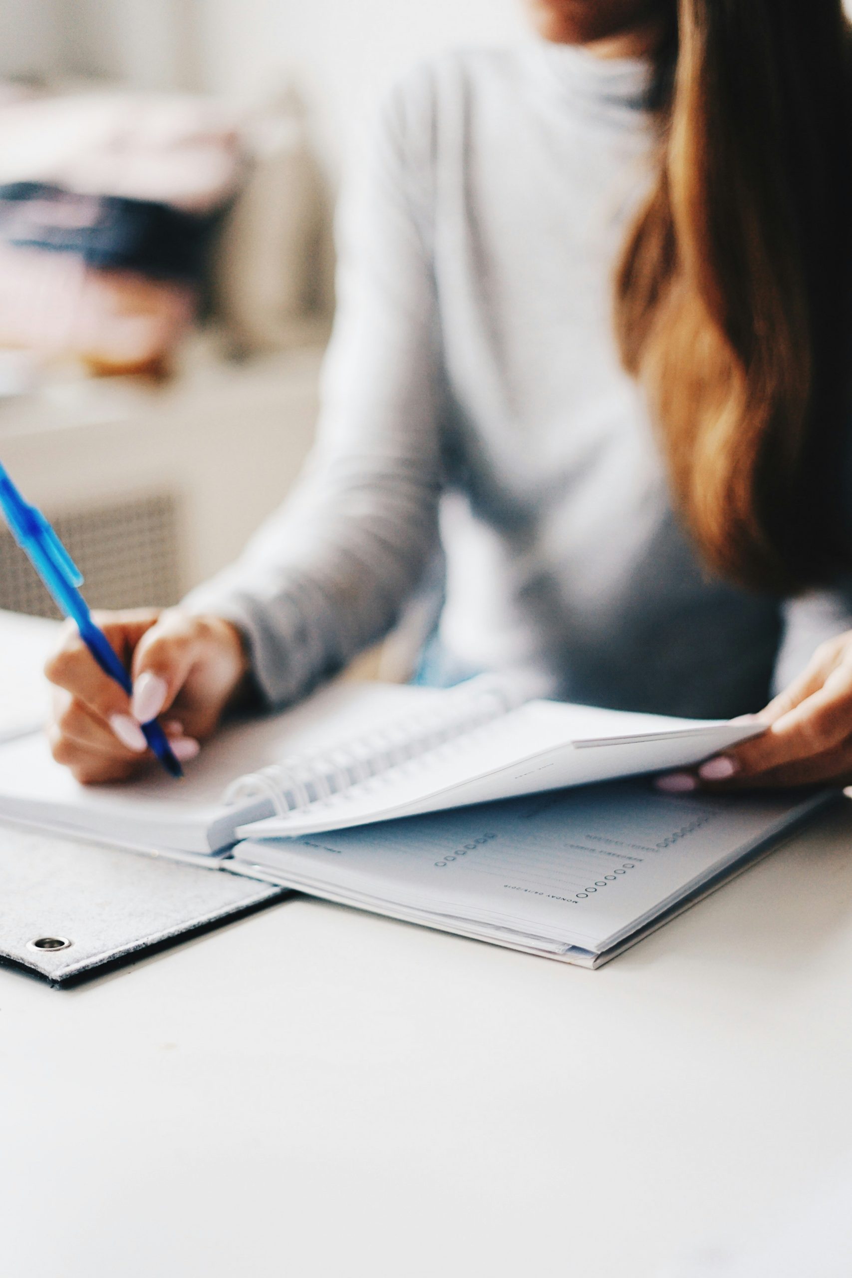 A person with long hair wearing a grey top writes in a notebook with a blue pen at a desk, with a planner and documents nearby.