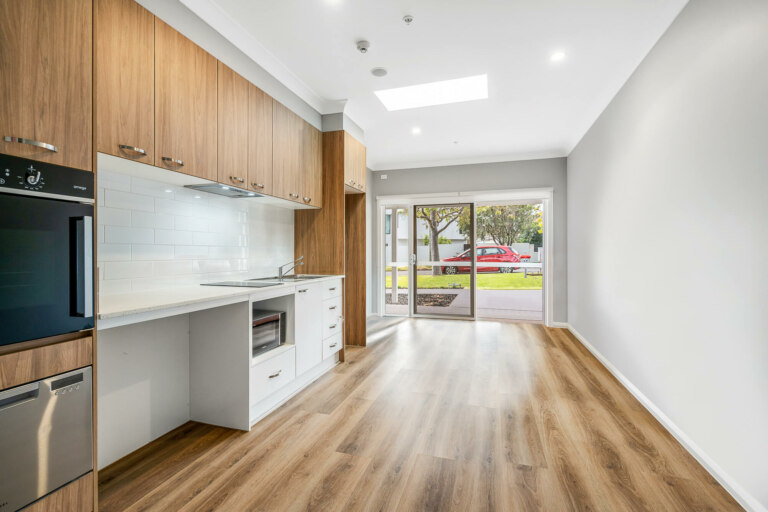 Modern kitchen with wooden cabinets, white countertops, built-in oven, and a dishwasher. An adjacent empty room in this Pagewood, NSW location features large windows overlooking a street with parked vehicles and greenery.