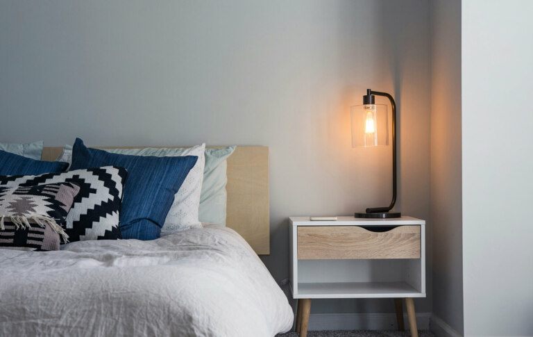 A modern bedroom in NSW, Maroubra features a comfortable bed illuminated by a sleek lamp and accompanied by a stylish bedside table.
