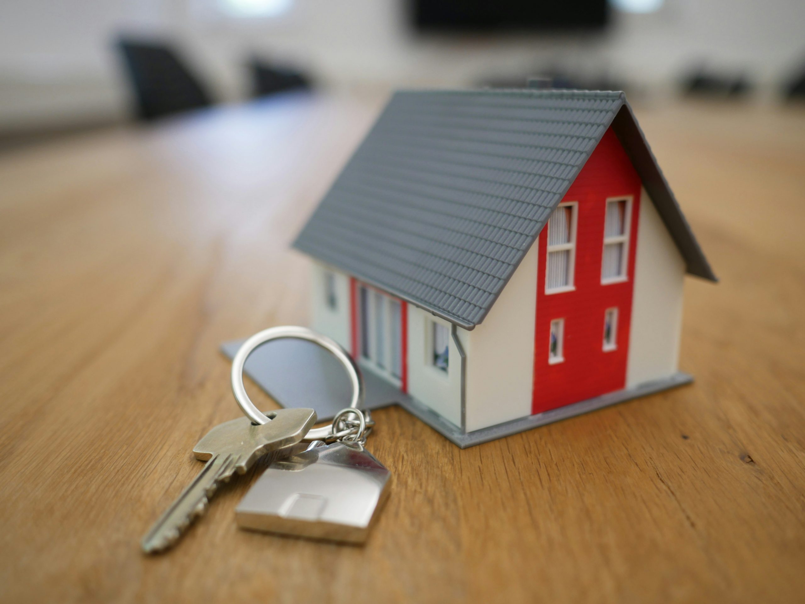A set of house keys on a keychain lies next to a small model of a red and white house on a wooden table.