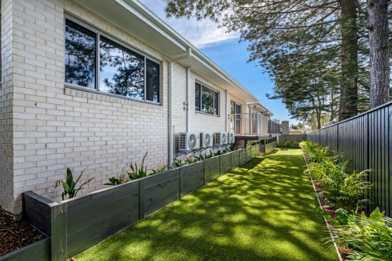 Single-story, white brick building with large windows and air conditioning units mounted on the side at 1A Narraweena. It has a landscaped sideyard with a neatly trimmed lawn, raised garden beds, and is flanked by a fence in Northern Beaches Sydney.