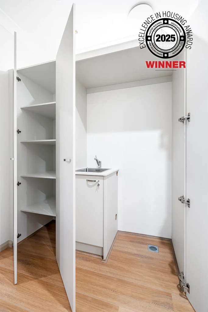 A small laundry area with open white cabinets, shelves, a sink, and a faucet on light wood flooring in Pendle Hill. A "2025 Excellence in Housing Awards Winner" badge is overlaid in the upper right corner.
