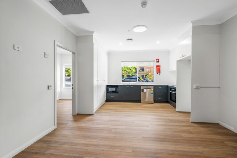 Experience modern living in this elegant kitchen located in a prime Sutherland NSW real estate. The space boasts wooden flooring, white walls, and a window that floods the room with natural light. Enjoy the convenience of a built-in oven, microwave, dishwasher, dark lower cabinets, and white upper cabinets.