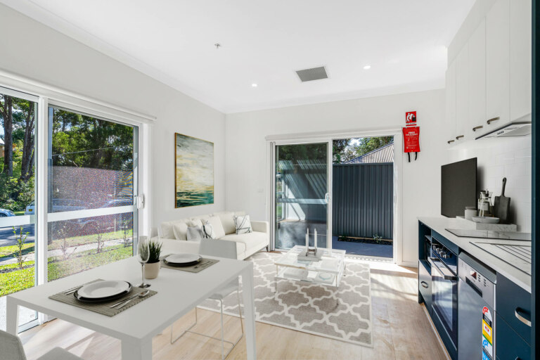A bright, modern kitchen and living area in Sutherland, NSW, featuring large windows, white cabinets, a small dining table, a couch, a wall-mounted TV, and a fire extinguisher on the wall.