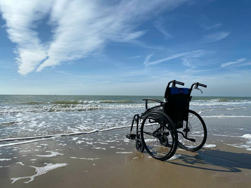 A wheelchair positioned on a sandy beach with foamy waves in the background under a blue sky with scattered clouds.